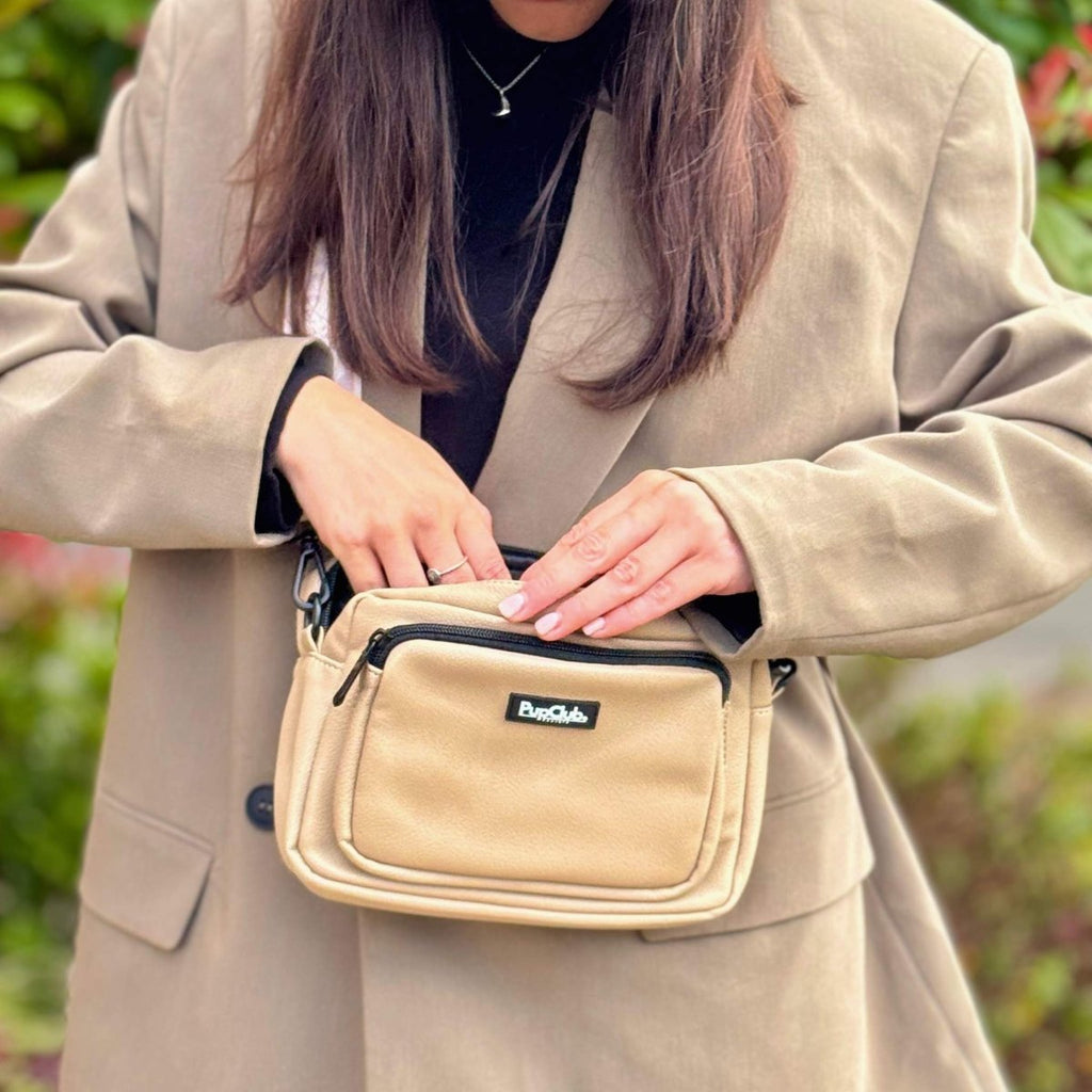 Tan crossbody bag with front zip pocket and PupClub logo patch, held at chest level