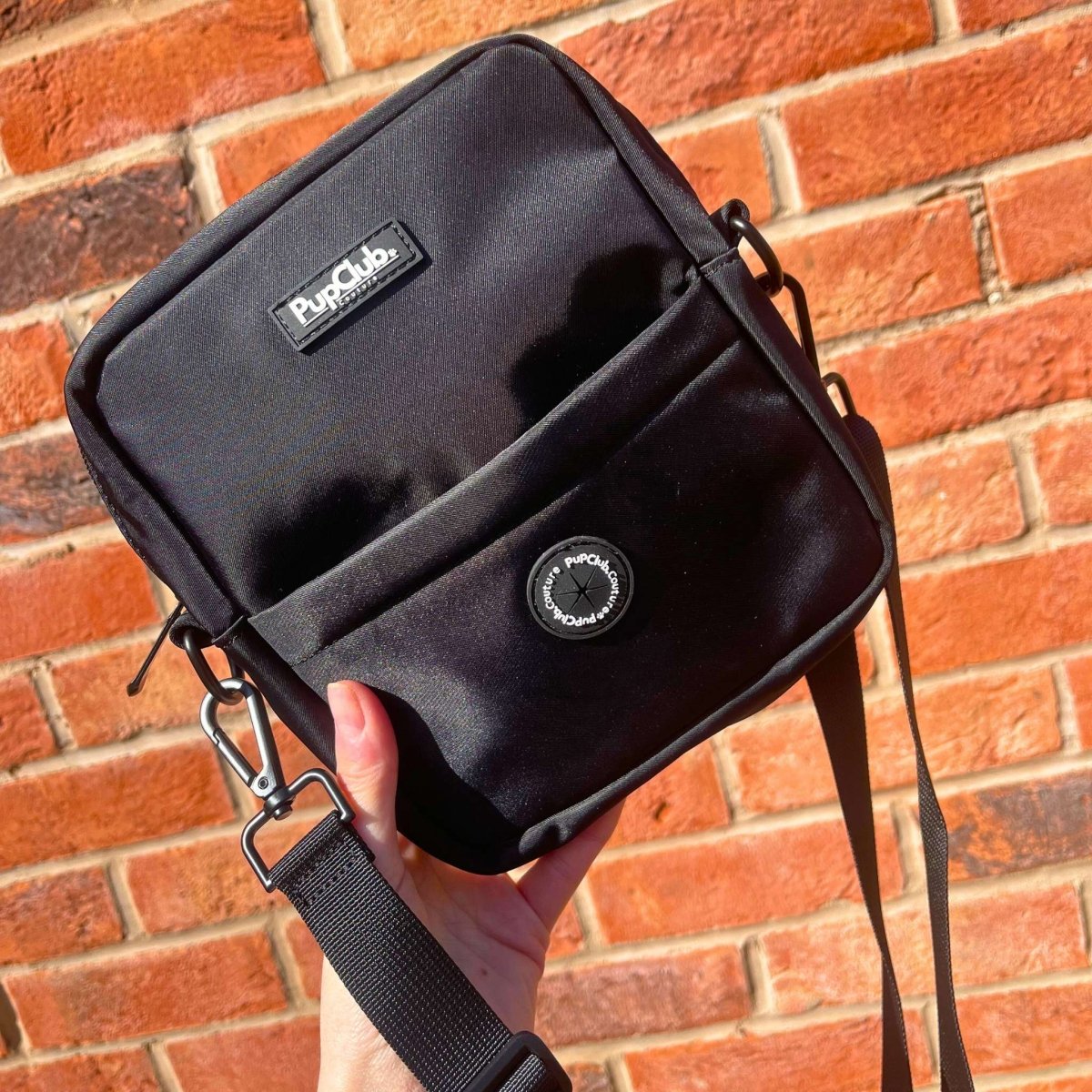 Black mini dog walking bag with front pocket, round badge, and carabiner strap loops